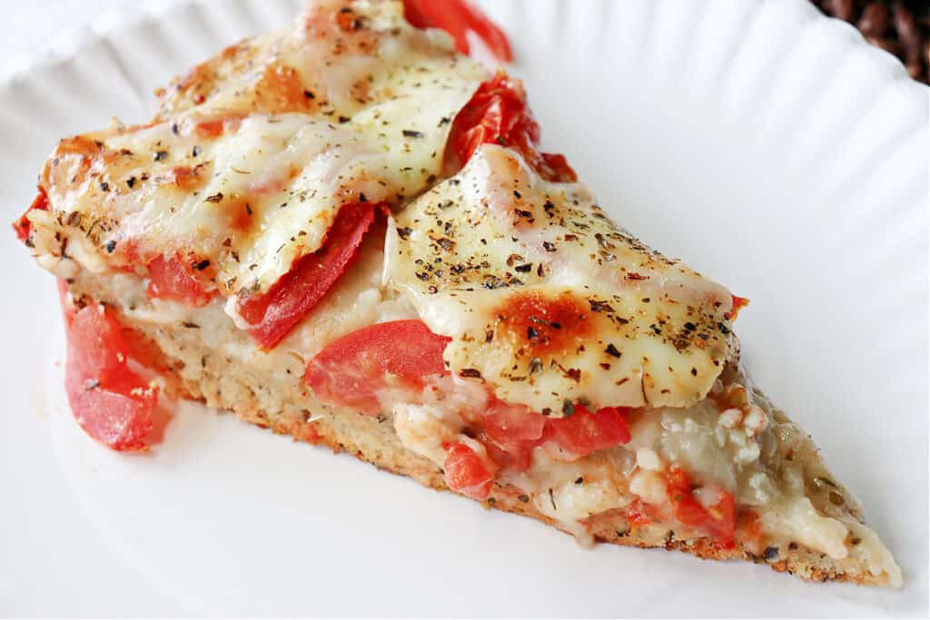 A closeup photo of a slice of Deep Dish Garlic Lover's Pizza on a paper plate with fresh tomatoes, melted cheese, and Italian seasonings.