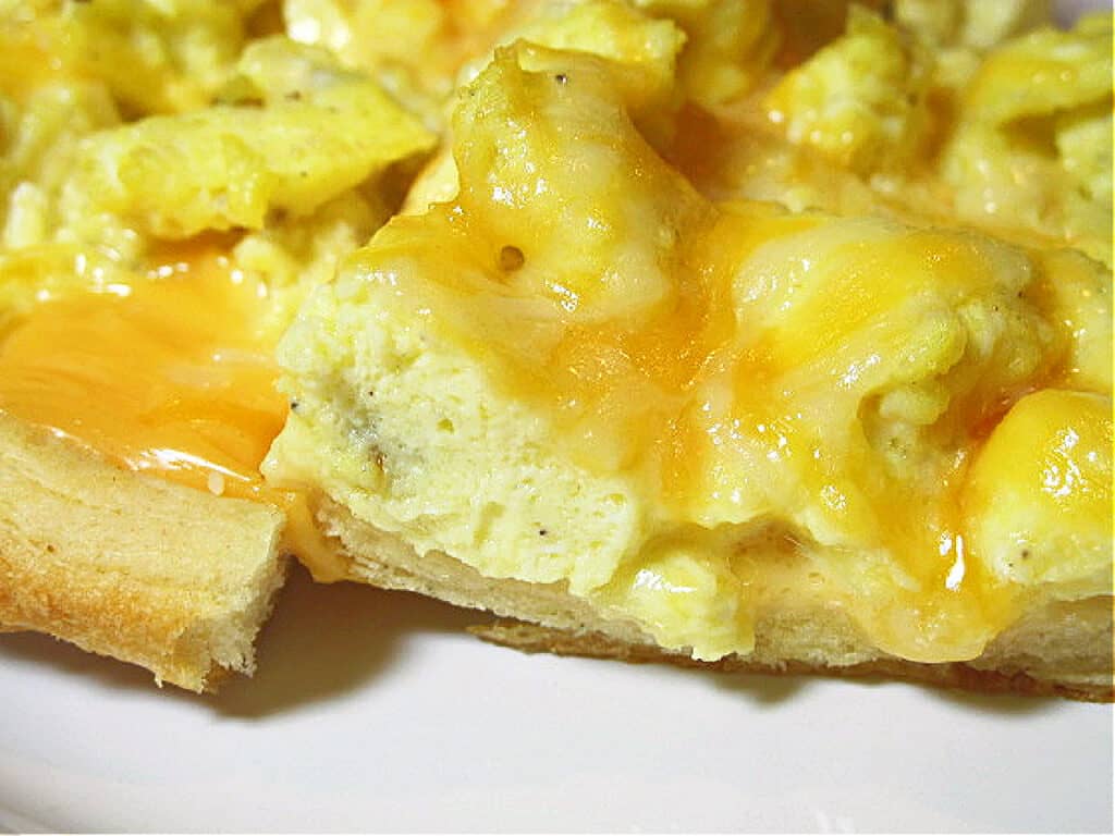 An extreme closeup of a slice of Scrambled Egg Breakfast Pizza with a golden brown crust and melted cheese on top.