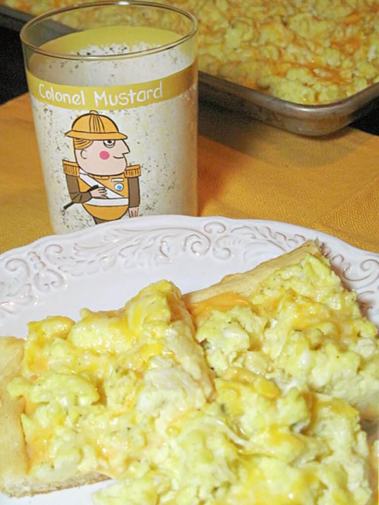 A vertical closeup of a few slices of Scrambled Egg Breakfast Pizza on a white plate with a yellow napkin and a painted glass in the background.