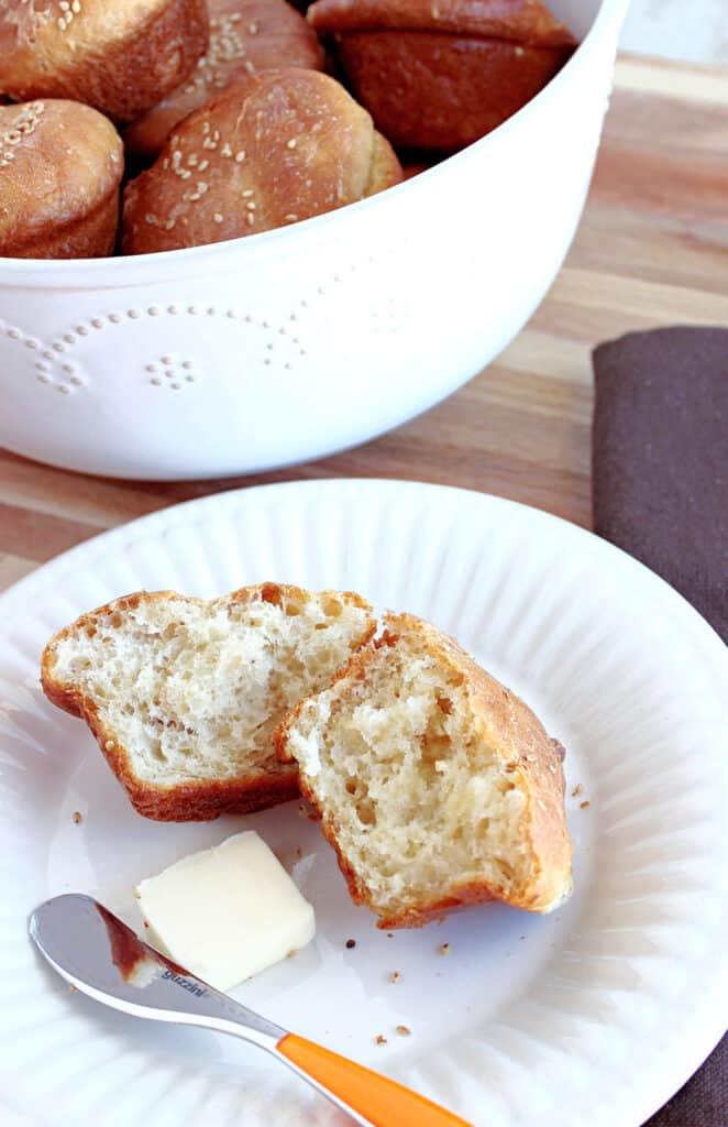 A vertical closeup of a bowl of Sesame Seed Dinner Rolls in the background and a white plate with a dinner roll and a pat of butter in the foreground.