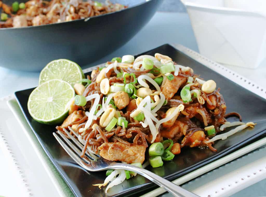 A serving of Chicken Pad Thai on a square black plate with lime wedges and a fork on the side.