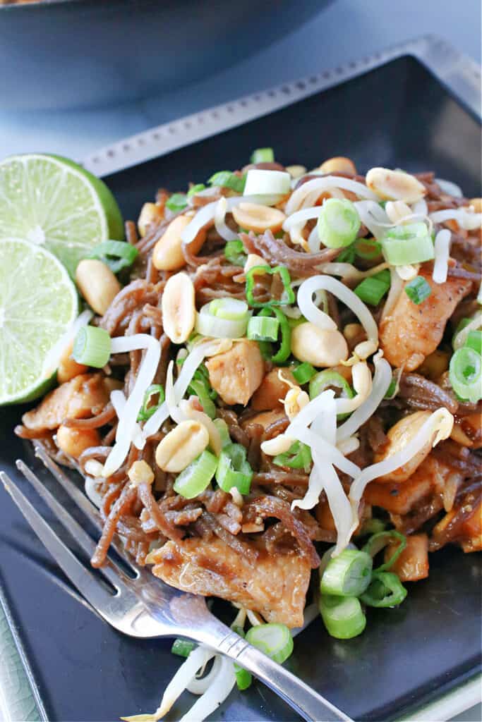 A closeup vertical image of Chicken Pad Thai on a black plate with a fork and limes.