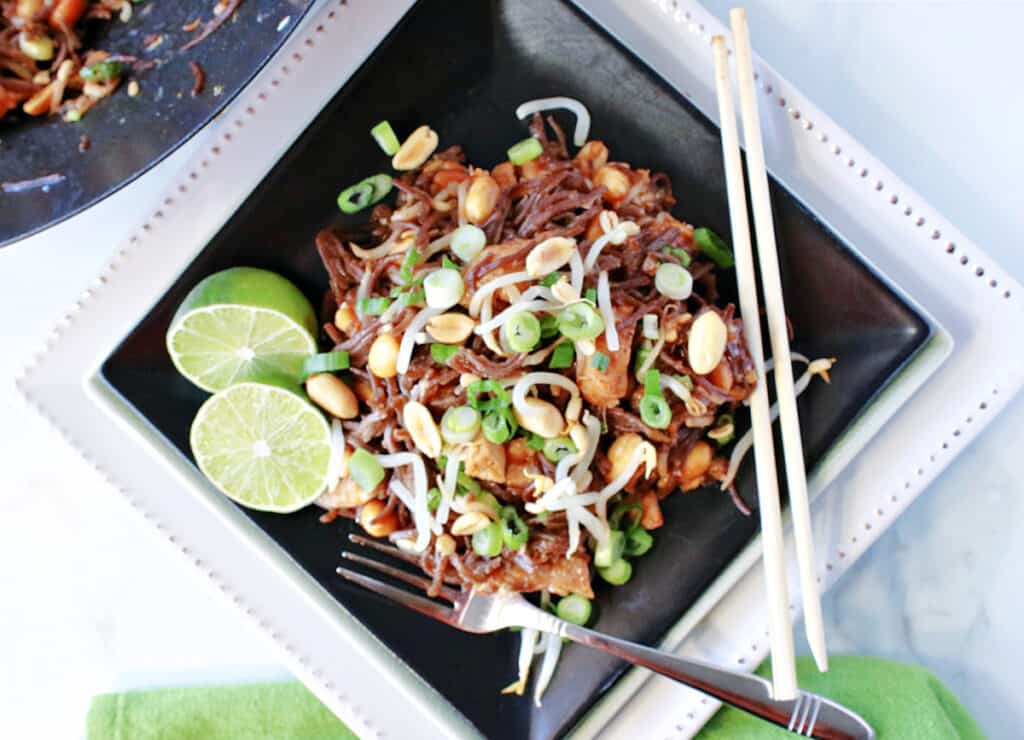 A direct overhead photo of a black square plate filled with Chicken Pad Thai along with limes, chopsticks, and a fork.