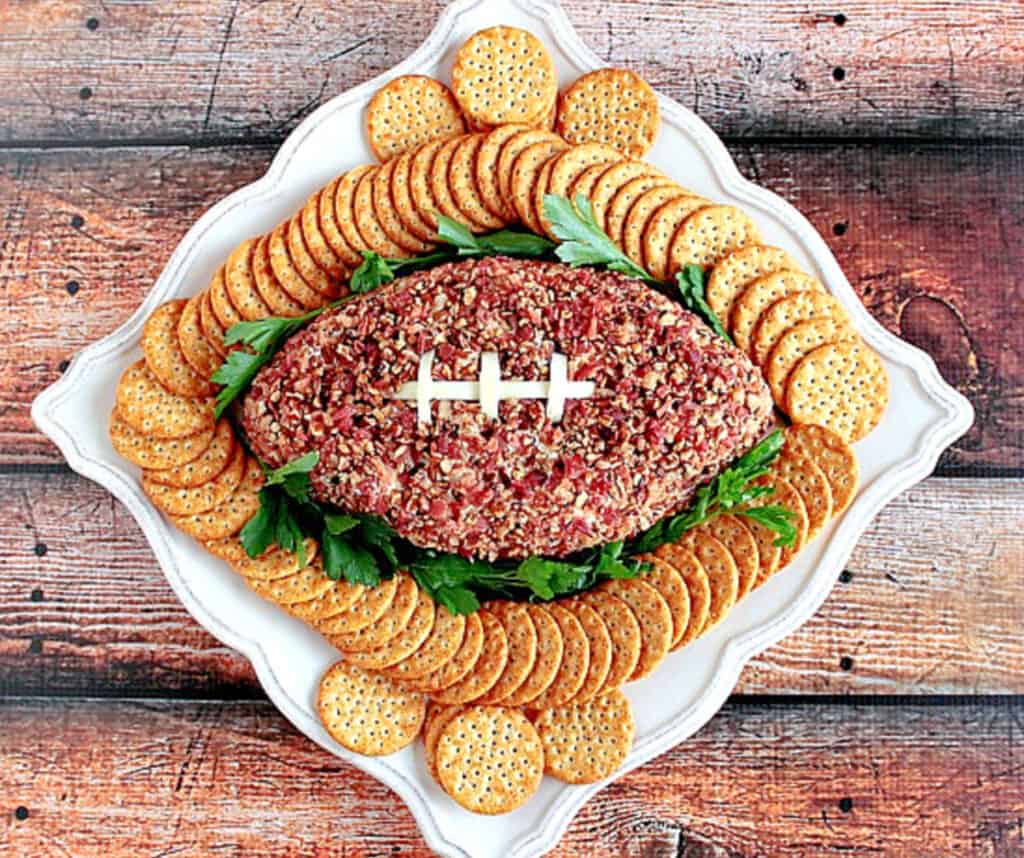 An overhead photo of a wooden table with a white platter filled with crackers and a Football Shaped Cheeseball in the center.