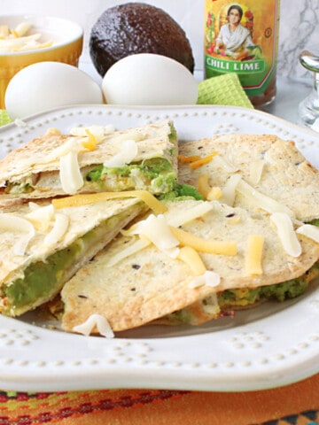 Triangles of an Grated Egg Avocado Quesadillas on a plate with a colorful napkin underneath and some avocados and eggs in the background.