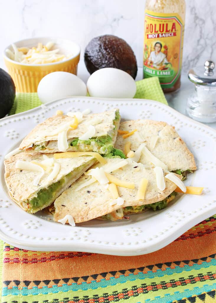 A vertical photo of a sliced Grated Egg Avocado Quesadilla on a plate with a bottle of hot sauce in the background.