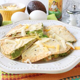Triangles of an Grated Egg Avocado Quesadillas on a plate with a colorful napkin underneath and some avocados and eggs in the background.