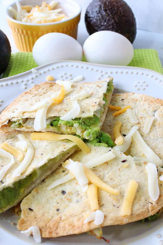 A closeup vertical image of a Grated Egg Avocado Quesadilla on a white plate with shredded cheese on top.