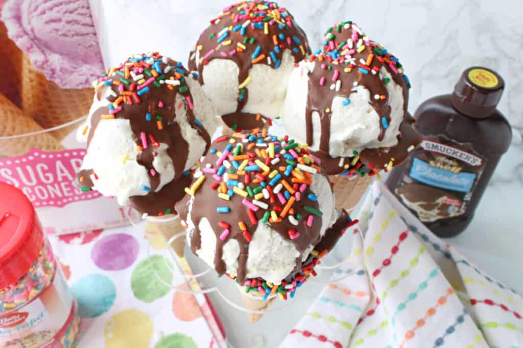 An overhead horizontal photo of four Ice Cream Cone Cake Pops topped with chocolate sauce and multi-colored sprinkles.