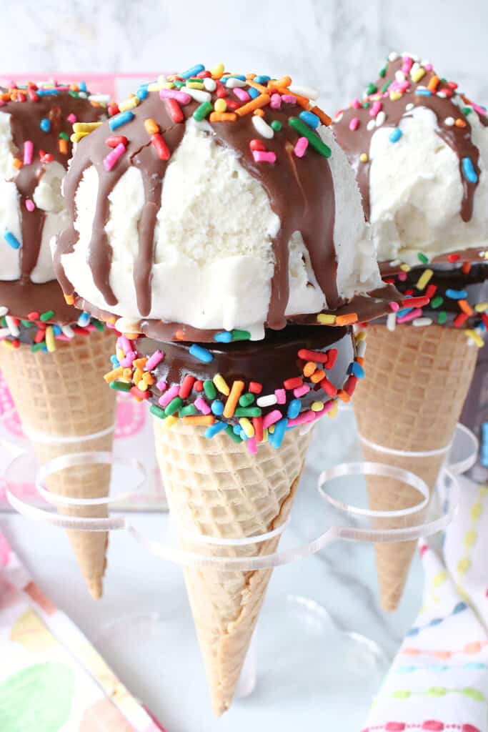 A vertical closeup of some Ice Cream Cake Pops in a plastic holder with chocolate sauce and sprinkles.