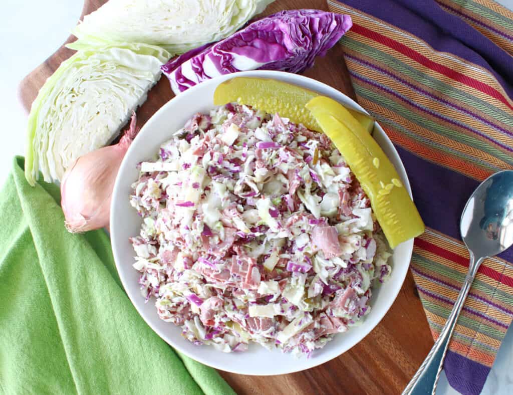 An overhead photo of a bowl of Corned Beef and Cabbage Coleslaw with a pickle on the side, some cabbage wedges, and a shallot.