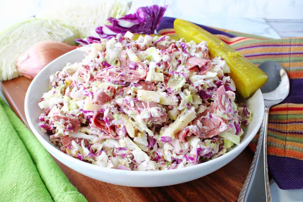 A white bowl filled with Corned Beef and Cabbage Coleslaw along with a pickle and cabbage wedges in the background.