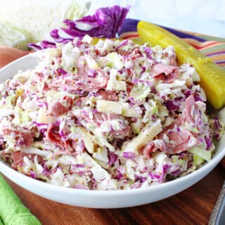 A white bowl filled with Corned Beef and Cabbage Coleslaw along with a pickle and cabbage wedges in the background.