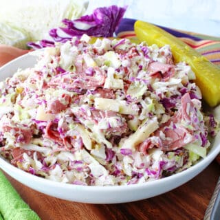 A white bowl filled with Corned Beef and Cabbage Coleslaw along with a pickle and cabbage wedges in the background.