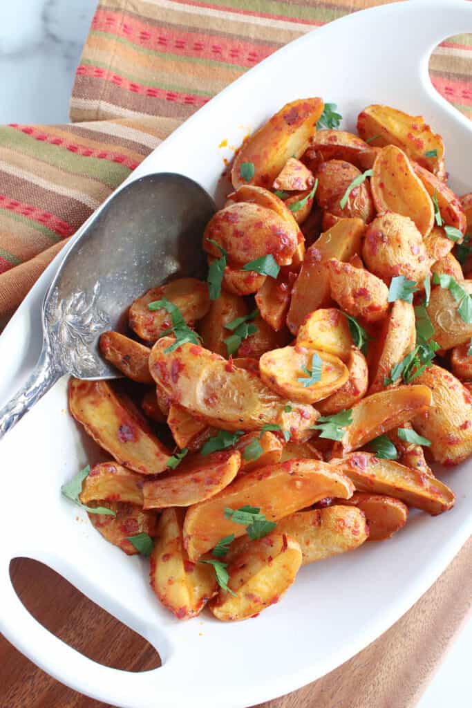 A vertical closeup of a bunch of Roasted Baby Potatoes with Chili Garlic Paste cut in half in a white bowl with a serving spoon and parsley.