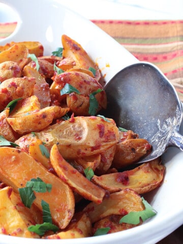 An offset white bowl filled with Roasted Baby Potatoes with Chili Garlic Paste along with a large serving spoon.