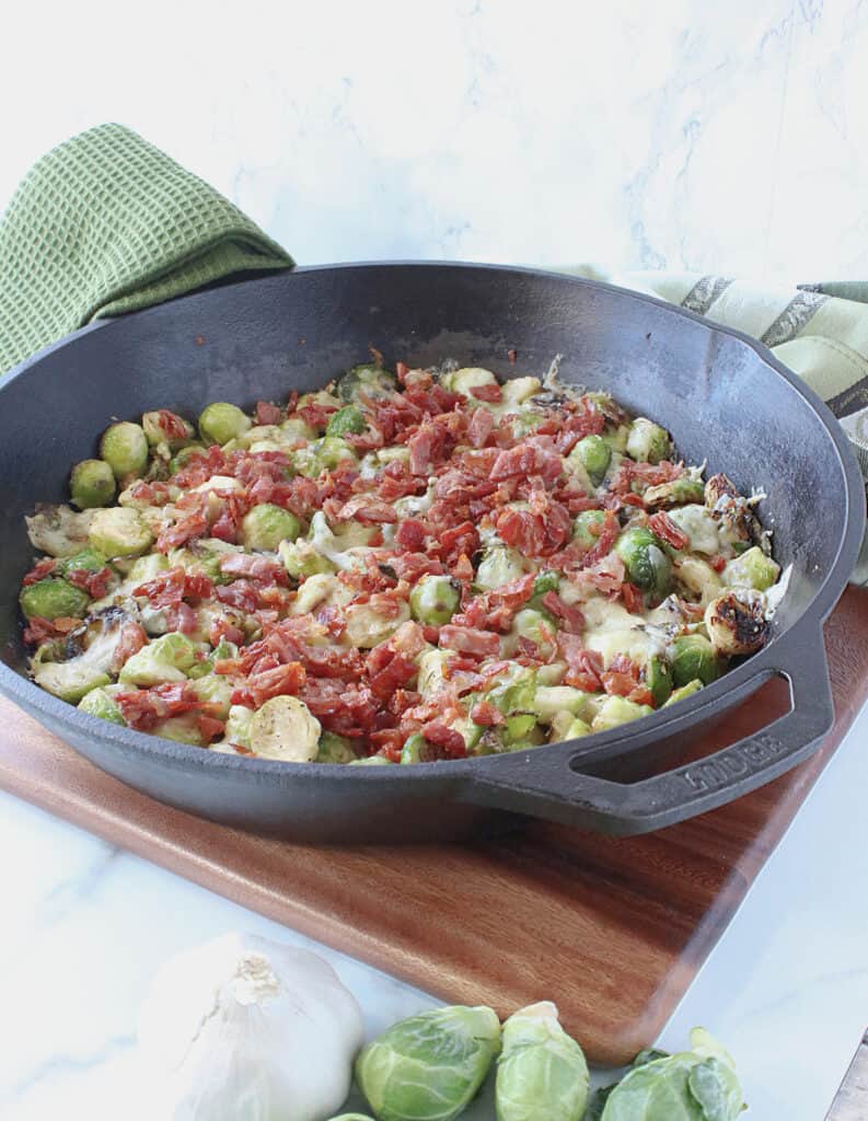 A vertical image of a cast iron skillet filled with cheesy Sautéed Brussels Sprouts with Crispy Prosciutto.