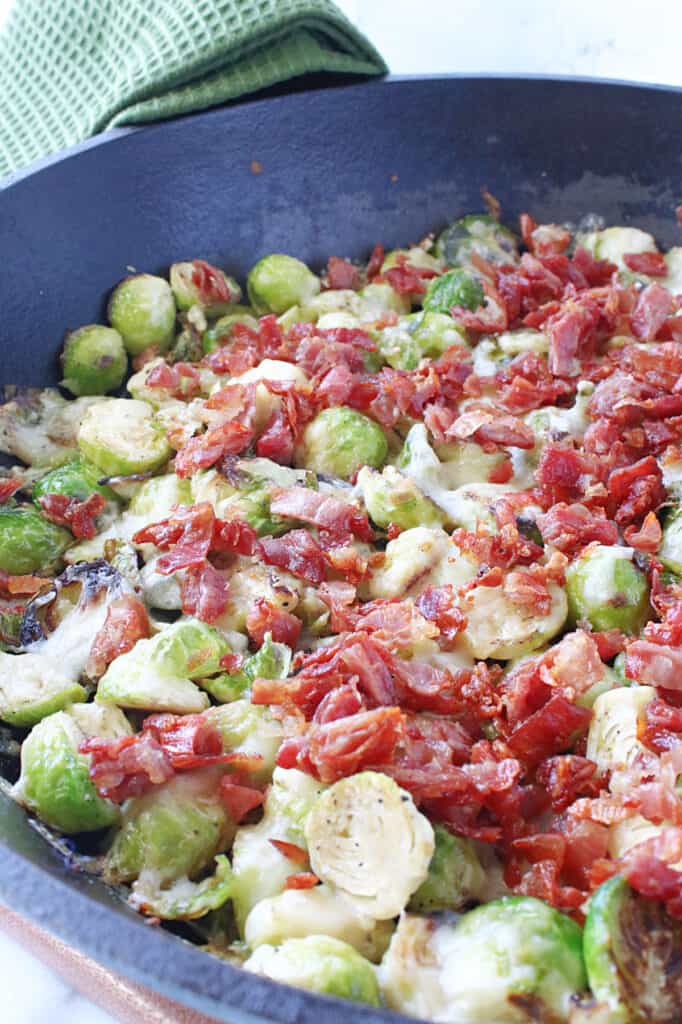 A vertical closeup of Sautéed Brussels Sprouts with Crispy Prosciutto in a cast iron skilled with melted cheese on top.