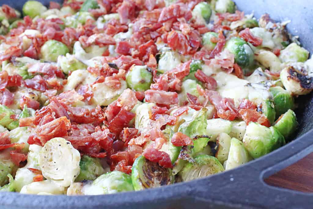 A closeup horizontal photo of Sautéed Brussels Sprouts with Crispy Prosciutto in a cast iron skillet with melted cheese on top.
