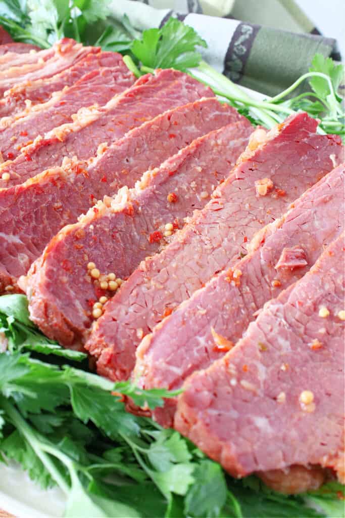 A vertical closeup photo of sliced Slow Cooker Corned Beef on a platter surrounded by fresh green parsley.
