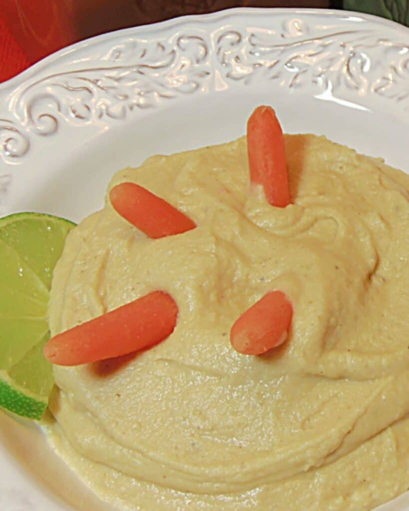 A super closeup photo of a bowl of creamy White Bean and Artichoke Hummus with baby carrots and a lime wedge.