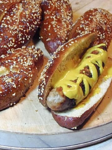 A group of Sausage Sized Pretzel Buns on a cutting board and one has mustard and a sausage inside.