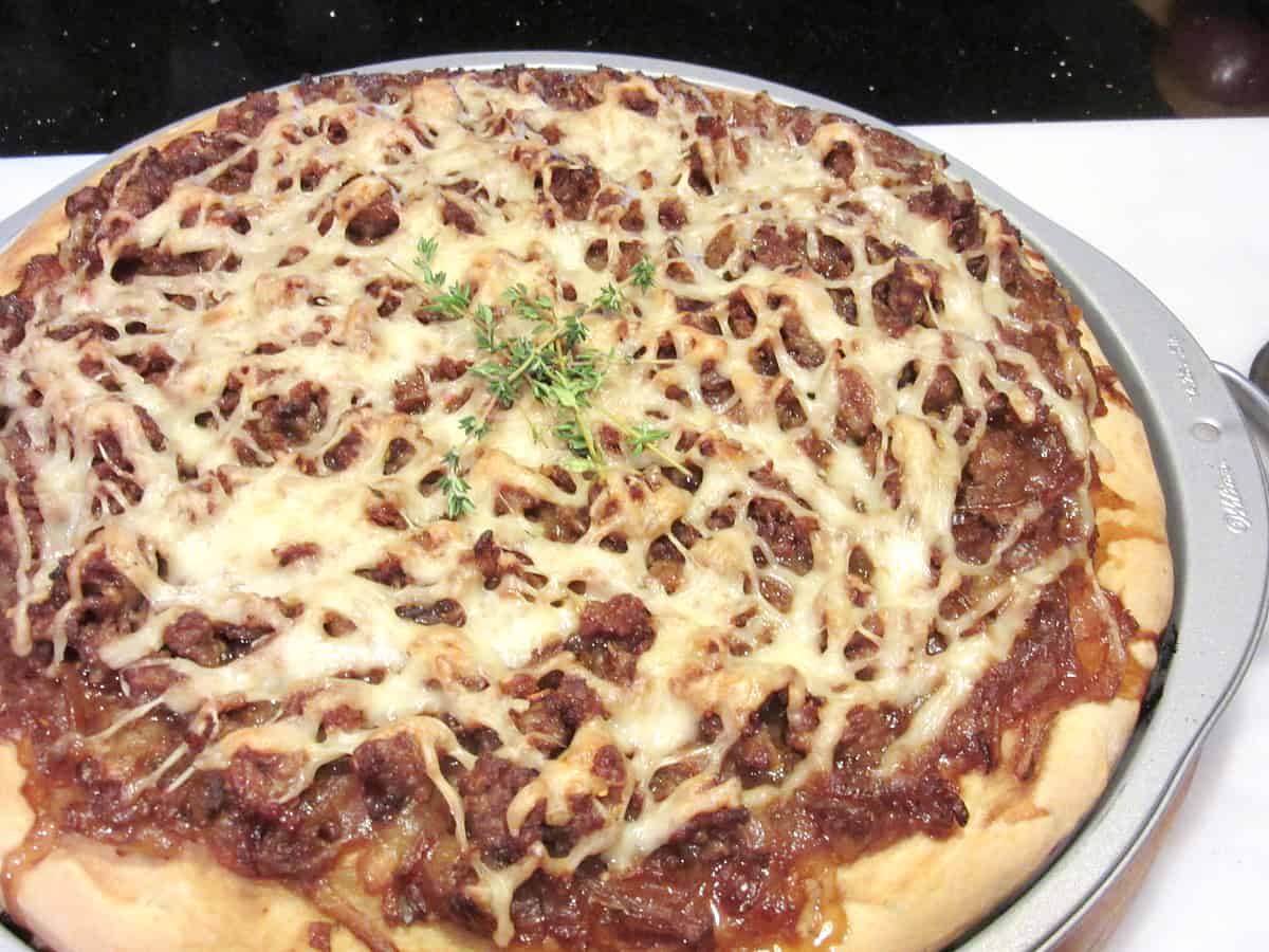 A French Onion Pizza on a pizza pan with melted cheese and fresh thyme leaves.