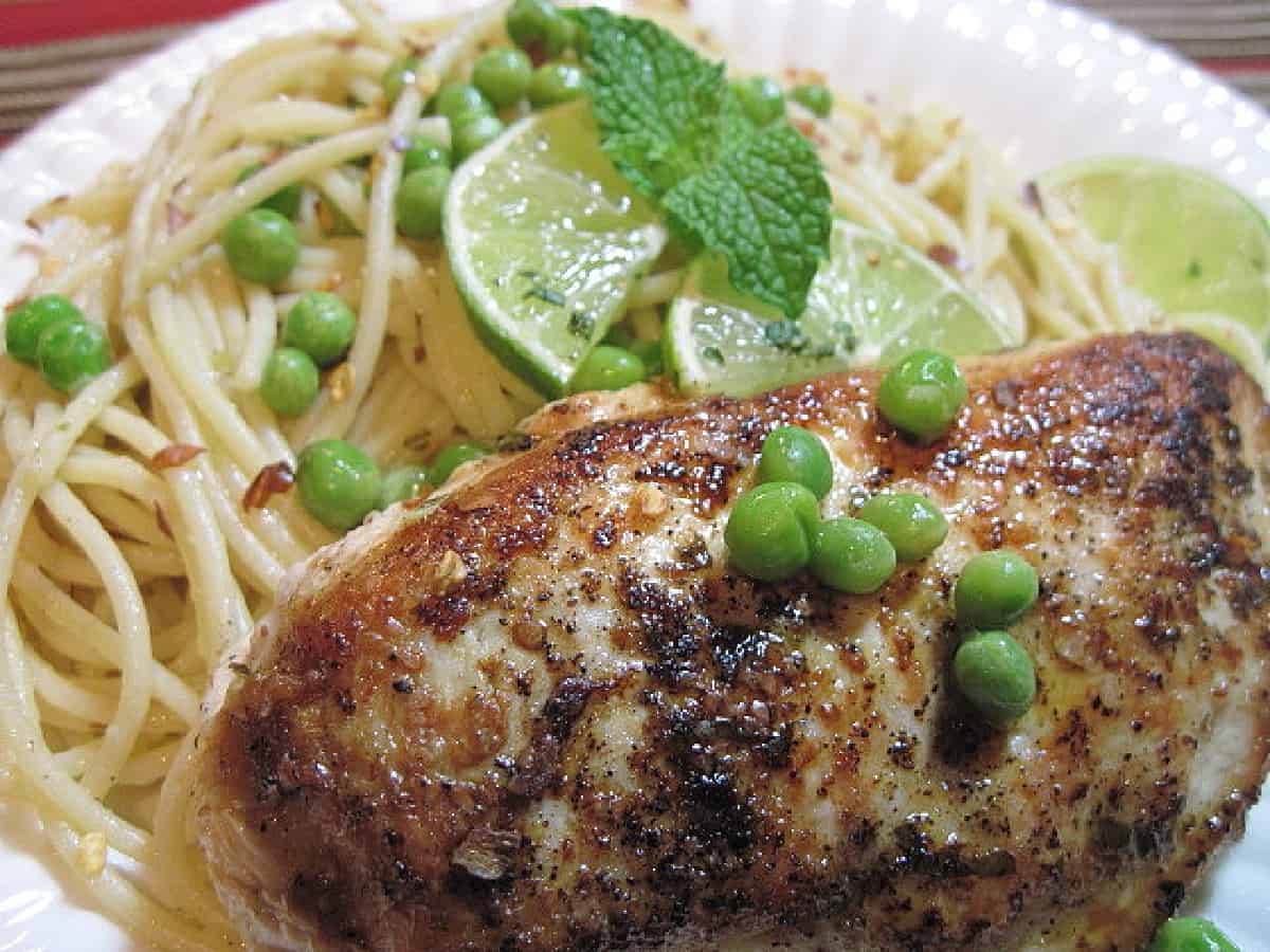A Mojito Chicken breast on a white plate with lime slices, mint, peas, and spaghetti.