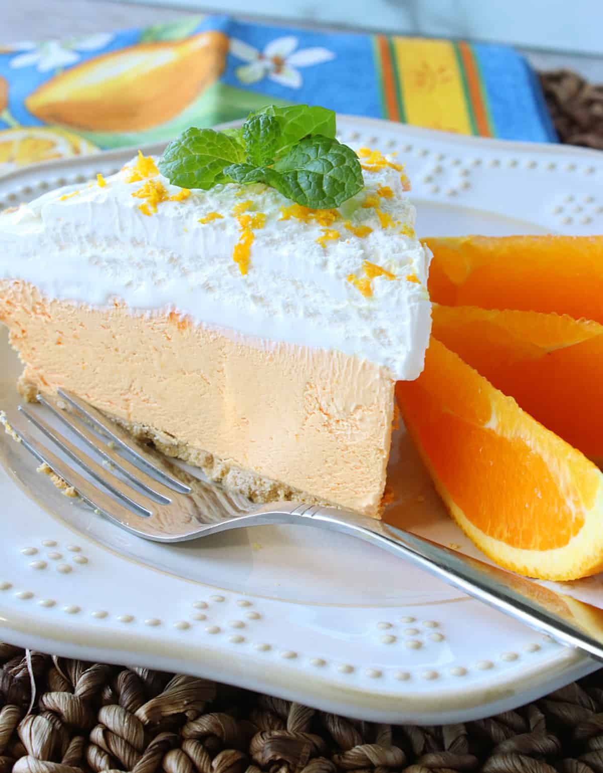 A vertical photo closeup of a slice of cream Cream Cheese Kool-Aid Pie on a plate with fresh orange slices on the side.