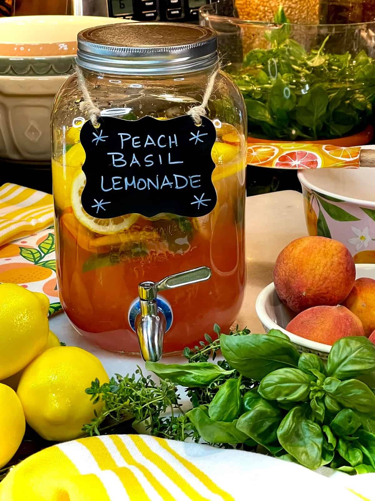A super colorful jug of Peach Basil Lemonade on the kitchen counter with fresh fruit and herbs all around.