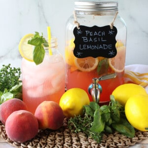 A glass and drink dispenser filled with Peach Basil Lemonade along with fresh fruit in the front.