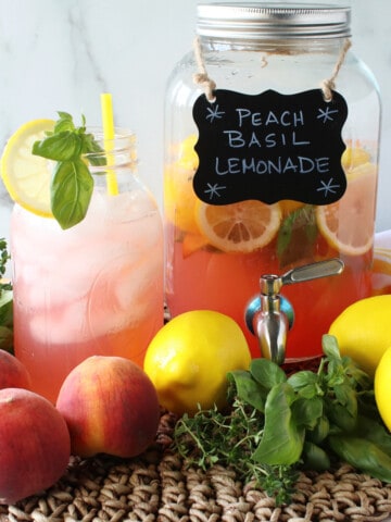 A glass and drink dispenser filled with Peach Basil Lemonade along with fresh fruit in the front.