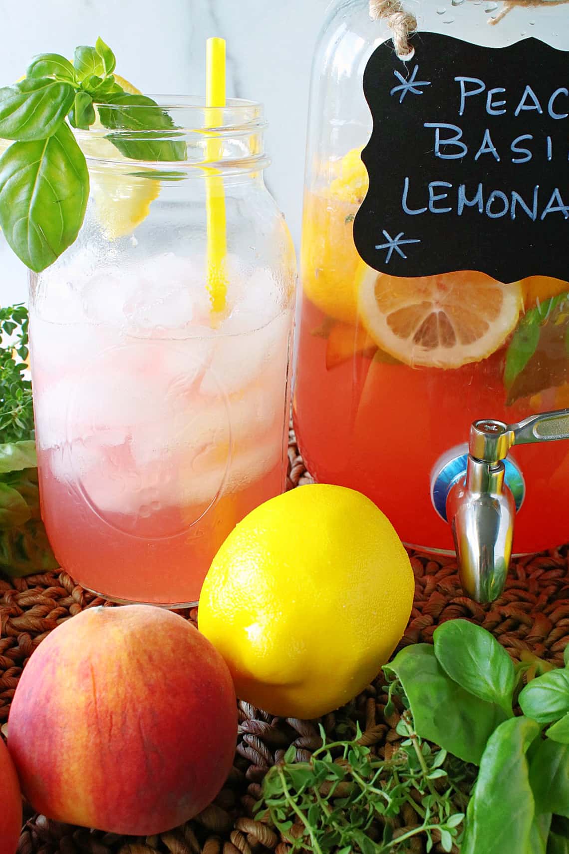 A glass mason jar with ice and a serving of Peach Basil Lemonade with a straw, lemon slice and fresh basil.