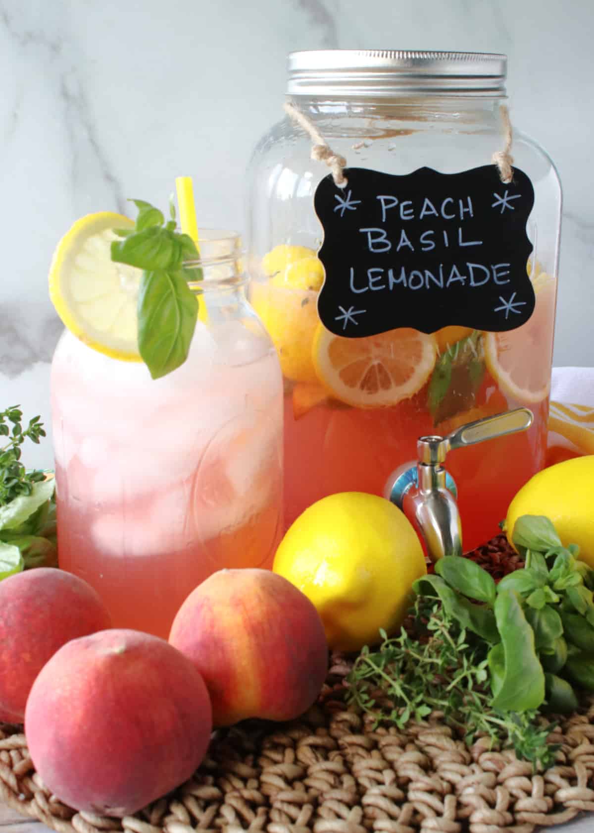 A glass pitcher with a spigot along with a glass with a straw of Peach Basil Lemonade.