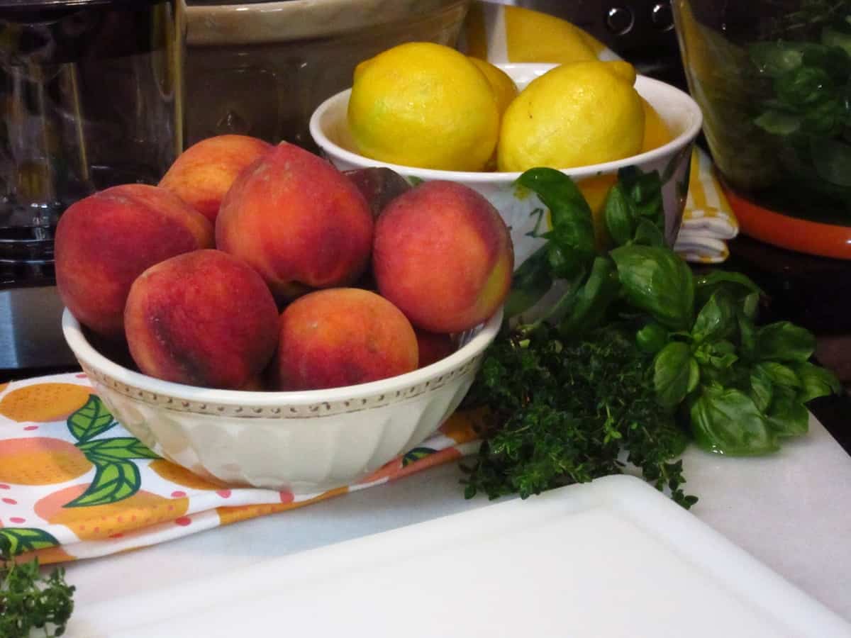 A bowl of peaches and lemons along with fresh basil and thyme on a counter.