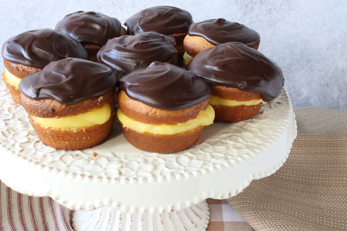 A pretty white floral cake plate topped with Boston Cream Cupcakes with chocolate ganache frosting.