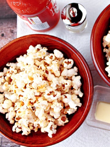 Two red bowls filled with Buffalo Popcorn along with a stick of butter and bottle of hot sauce in the background.