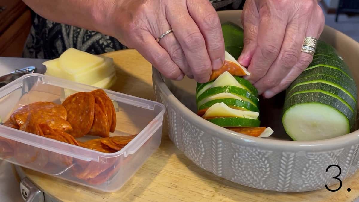 A pair of hands adding cheese and pepperoni slices to a Hasselback Zucchini.