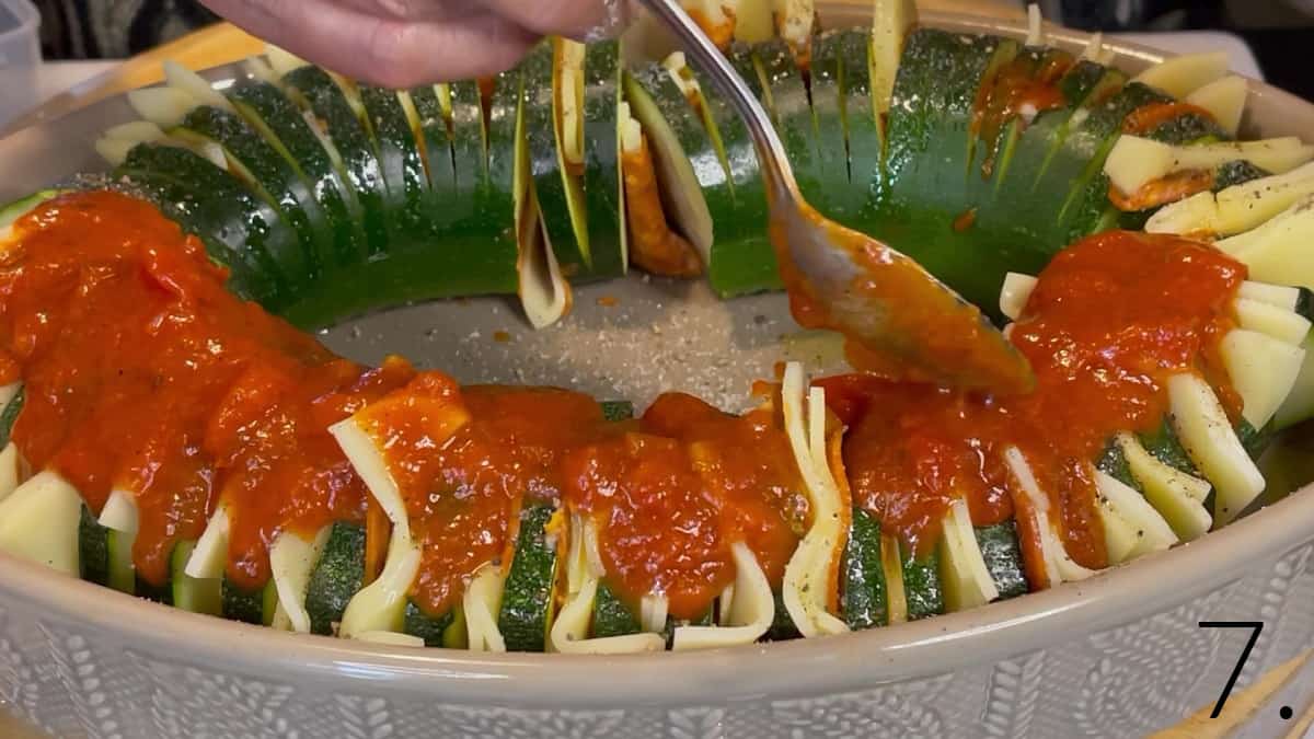 Marinara sauce being spooned over a unbaked Hasselback Zucchini with cheese and pepperoni.