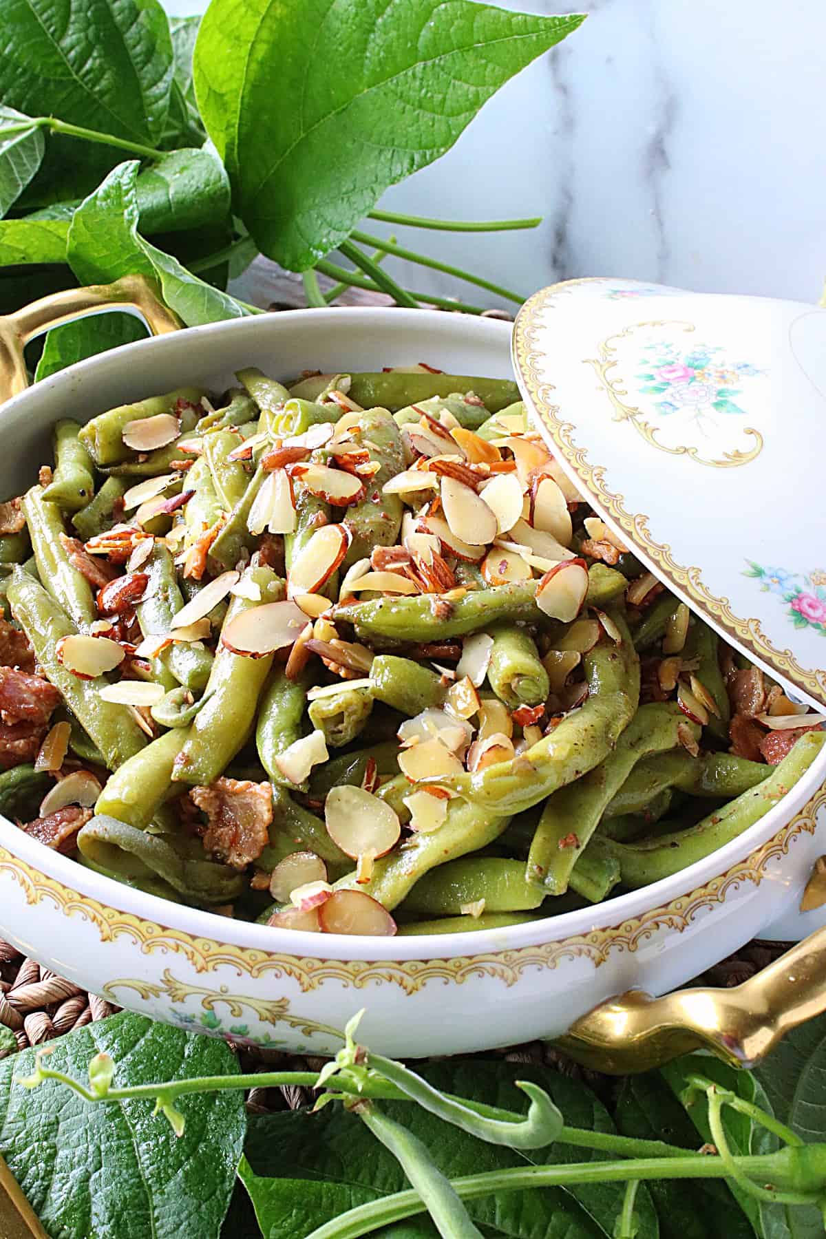 A pretty china bowl filled with Bacon Bourbon Green Beans Almondine with green bean leaves in the foreground.