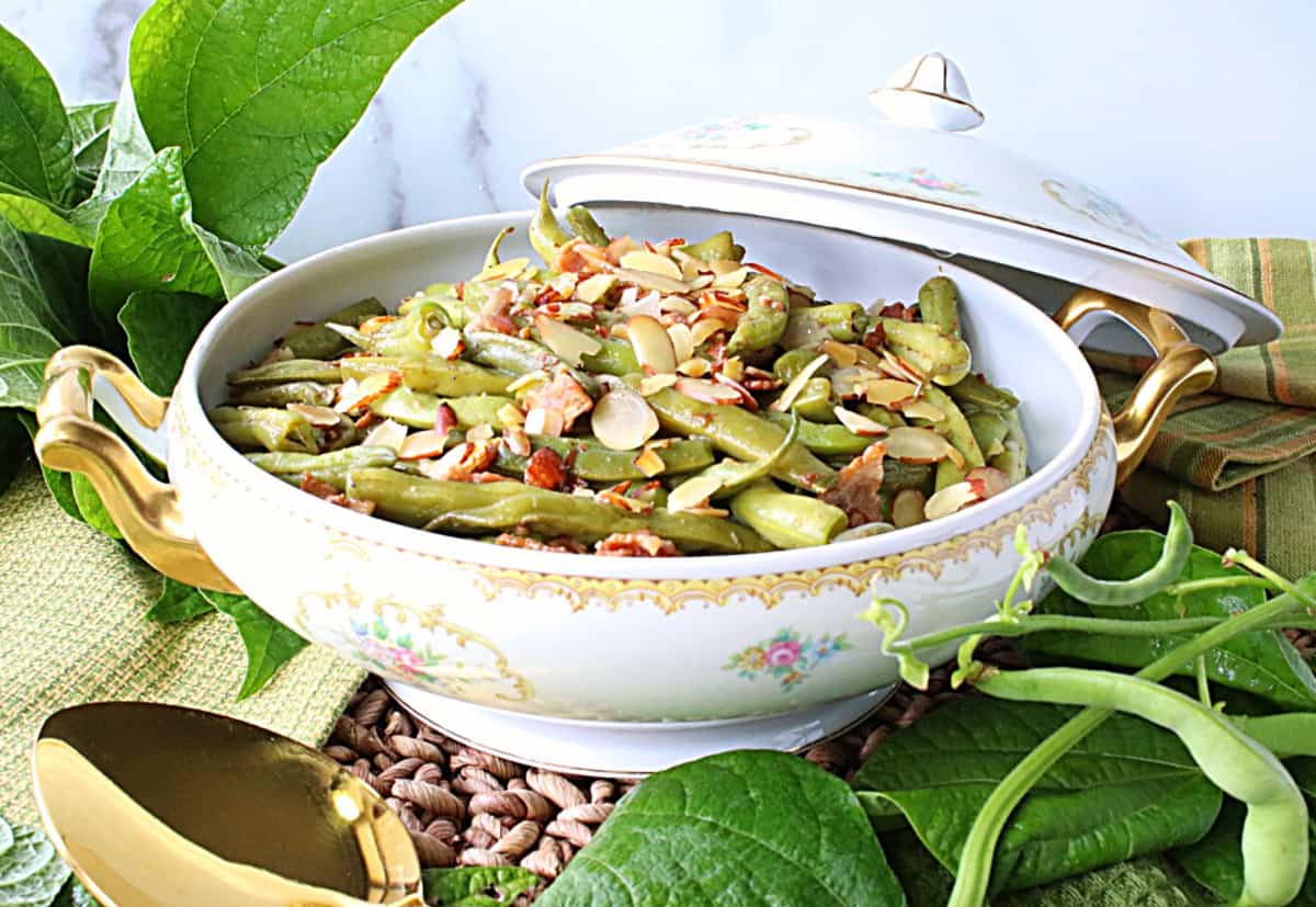 A delicious looking bowl of Bacon Bourbon Green Beans Almondine along with fresh leaves and beans as accents.