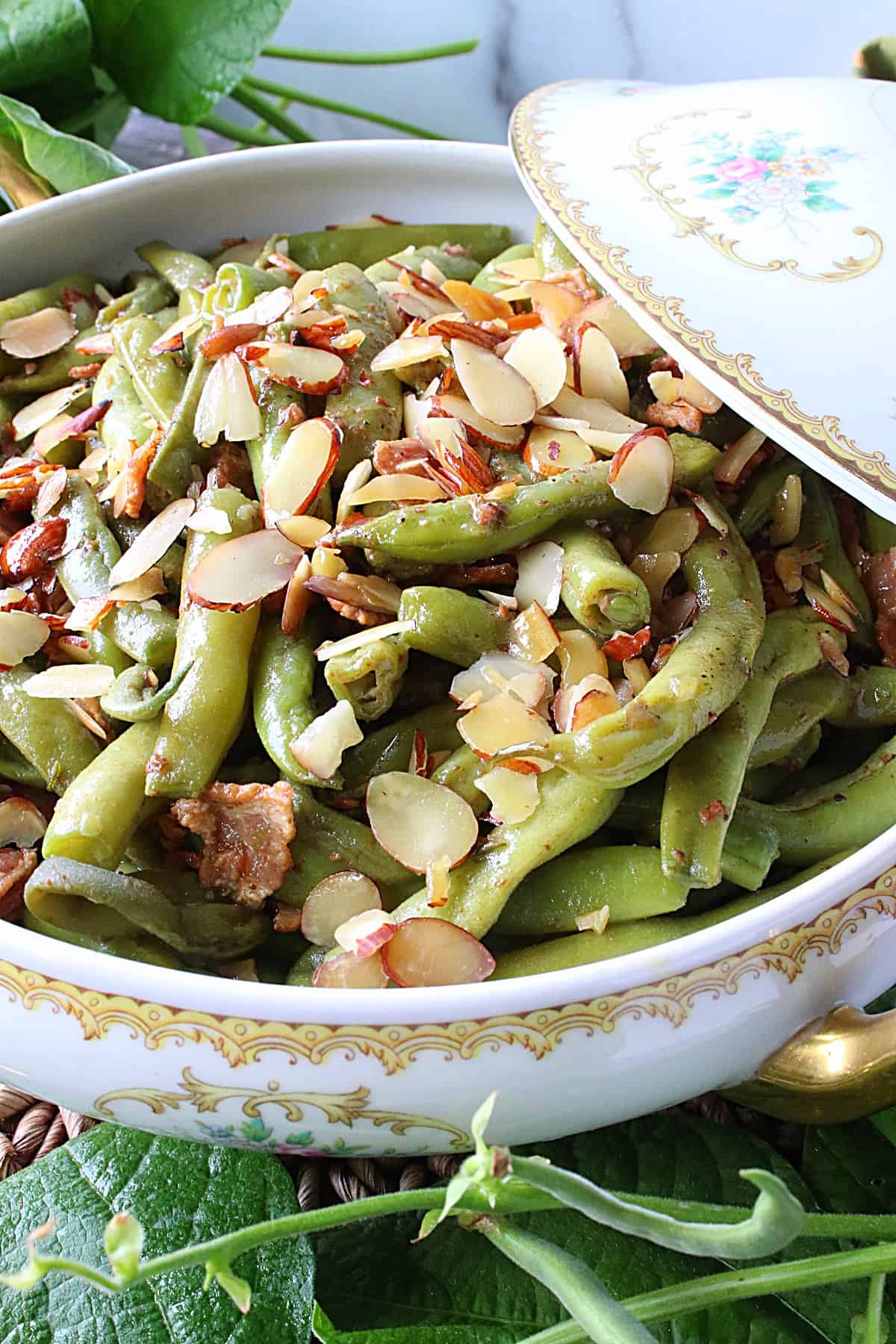 A closeup of a china serving bowl with a lid filled with Bacon Bourbon Green Beans Almondine.