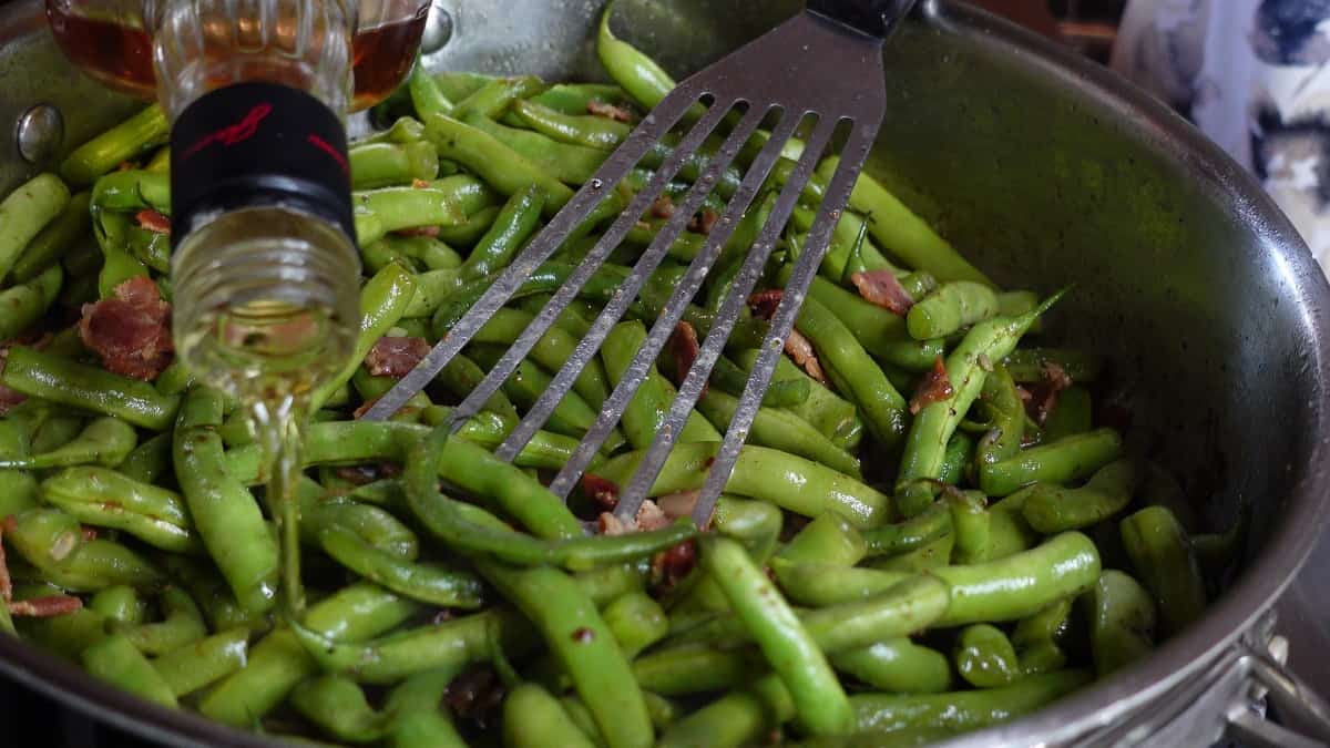 Adding bourbon to a skillet filled with green beans and bacon.