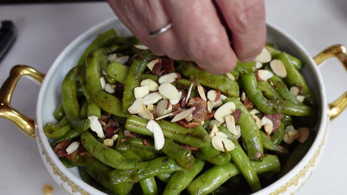Adding toasted sliced almonds to a serving bowl of bacon bourbon green beans almondine.