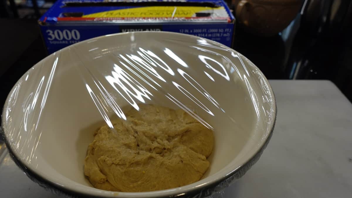 A mixing bowl with dough covered with plastic wrap for proofing.
