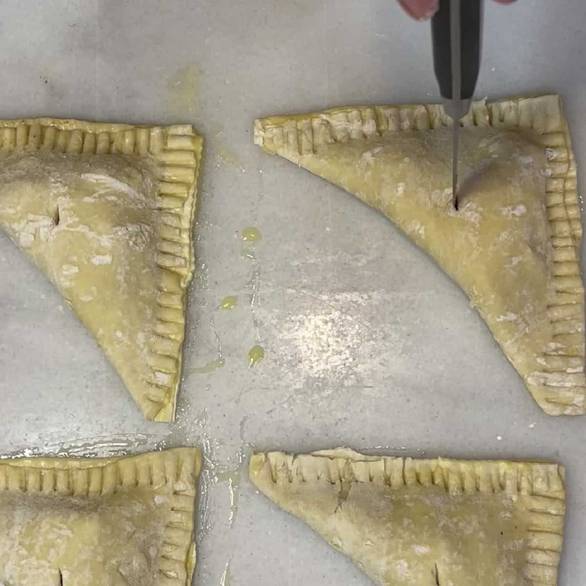 A paring knife cutting steam holes on top of unbaked apple turnovers.