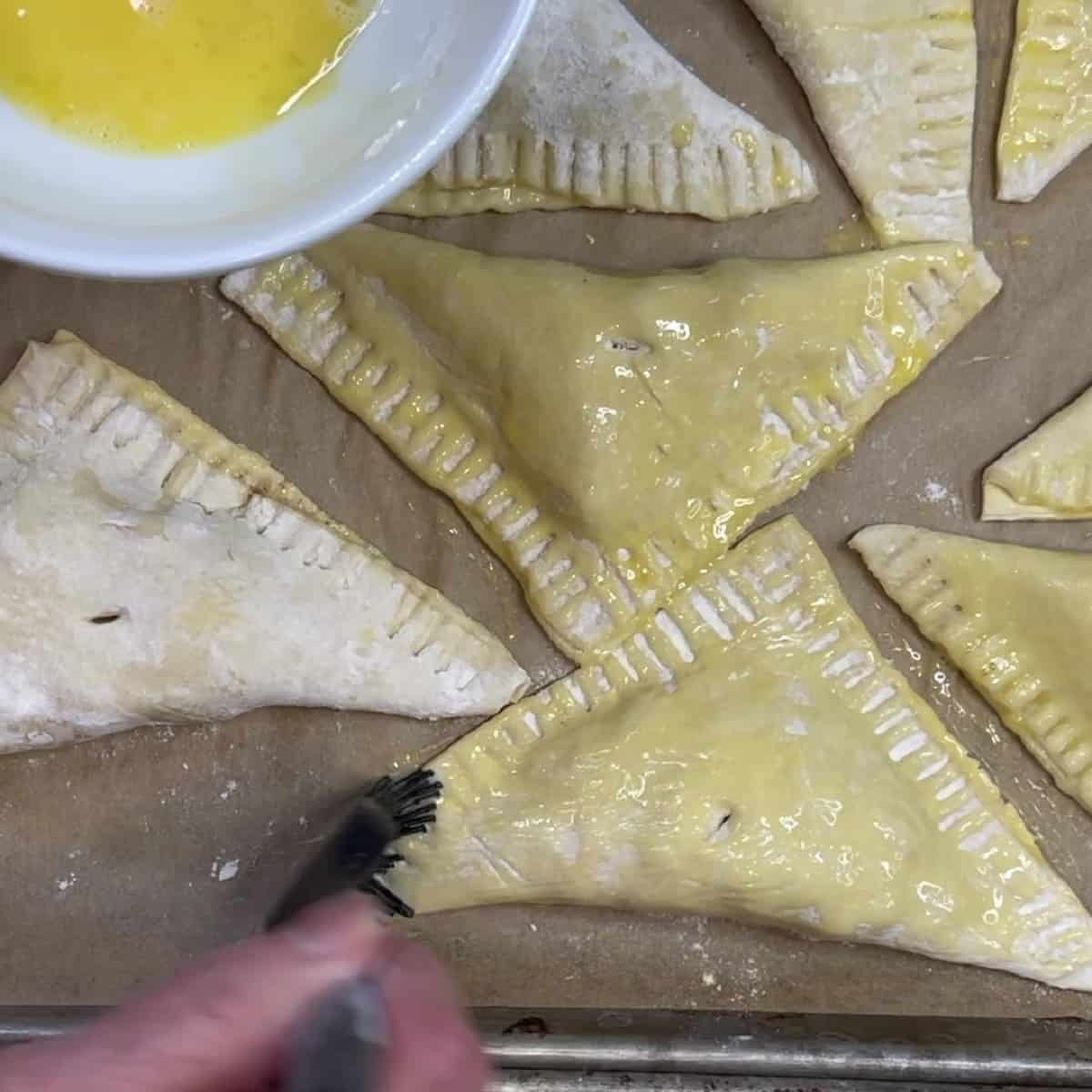 Egg wash being brush on top of unbaked puff pastry apple turnovers.