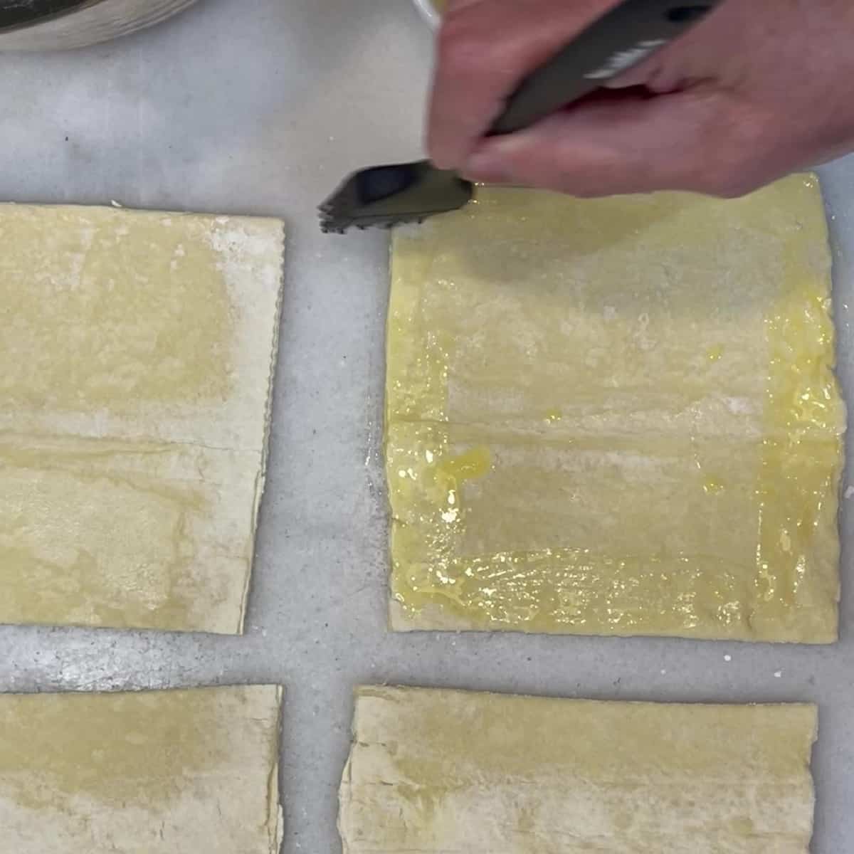 A pastry brush brushing egg wash onto puff pastry squares.