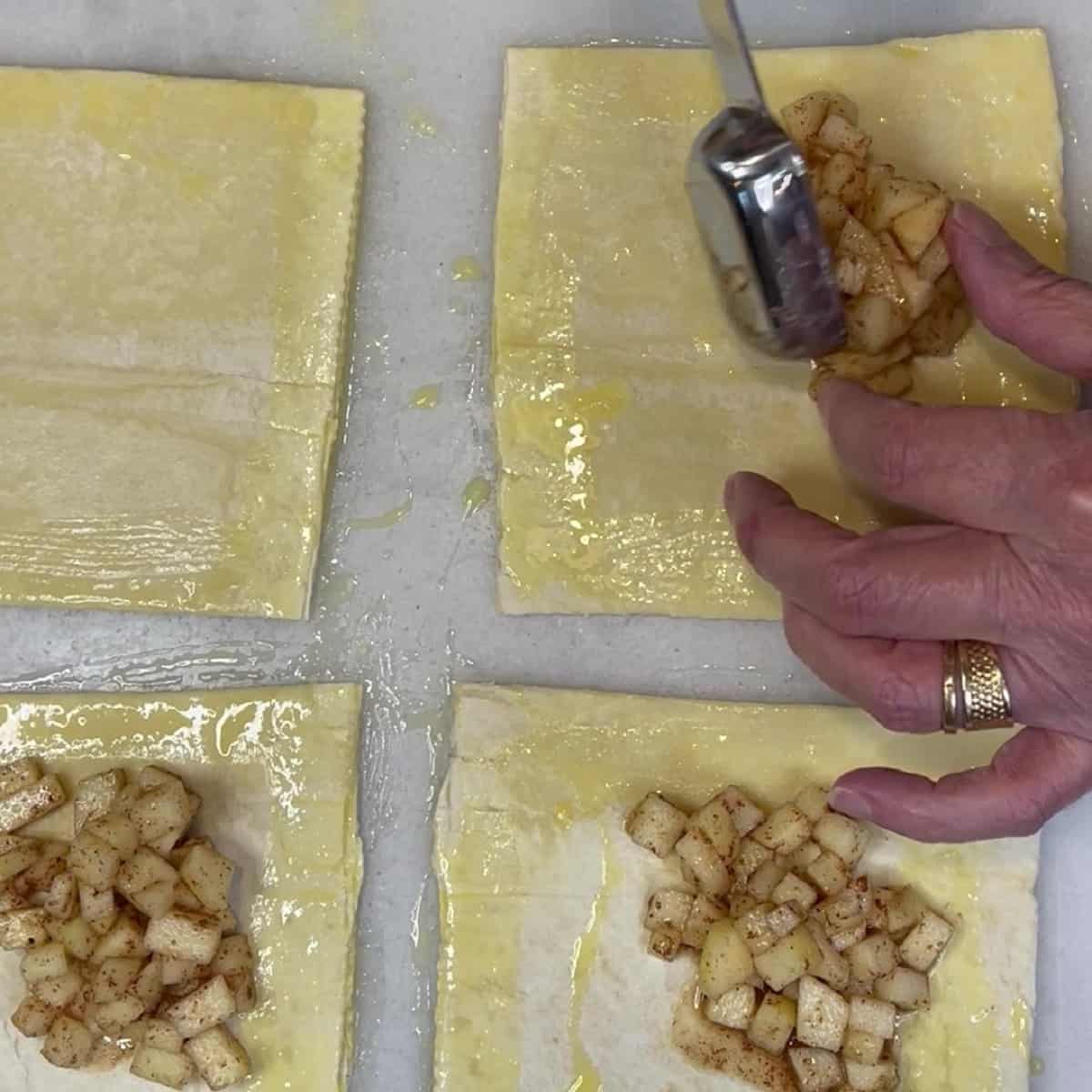 Diced apples being placed onto puff pastry squares to make turnovers.