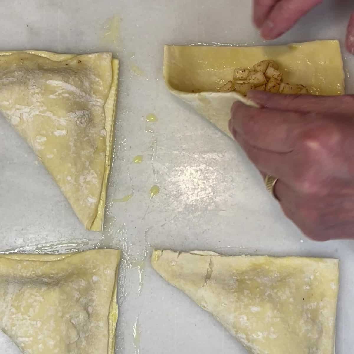 A hand folding over the puff pastry to make an apple turnover.